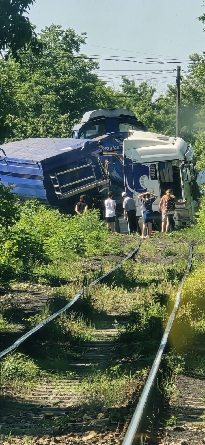 Accident la Motru între un tren de marfă și un camion Sursa Foto : FB/Anamaria Mitroi - Jurnalul Olteniei – Știri din Craiova și Oltenia Blue and white freight train derailed on overgrown tracks with a group of people inspecting the wreckage nearby.