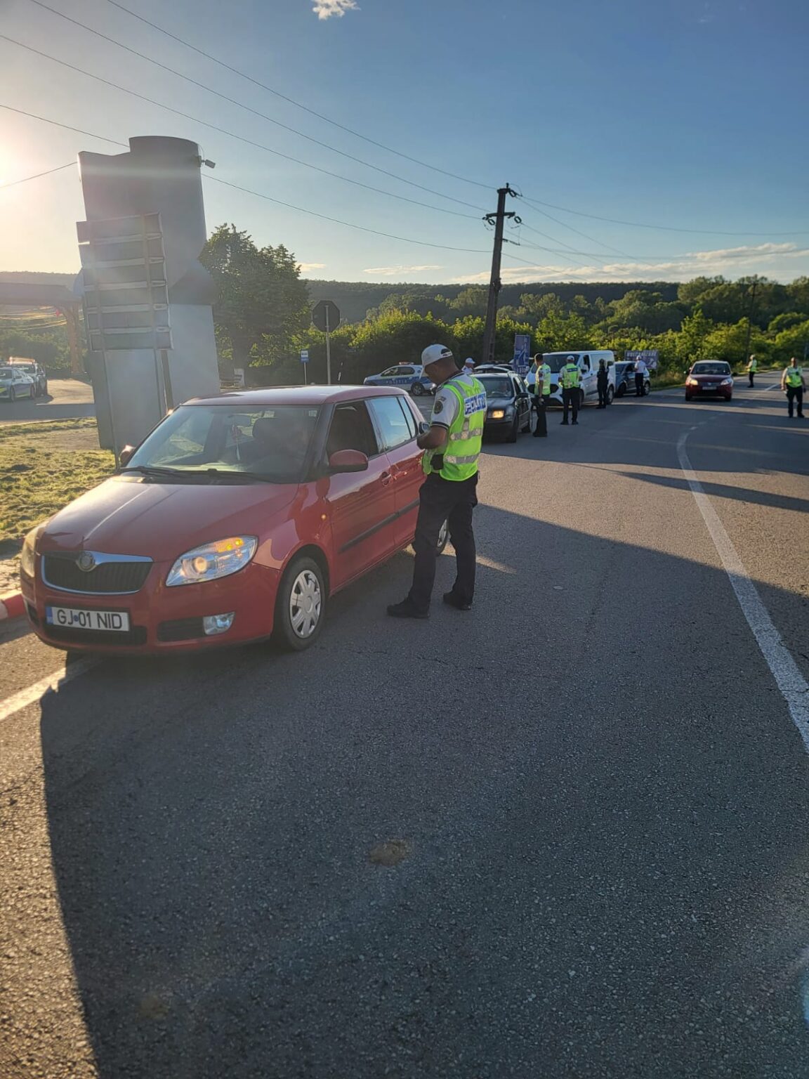 Police officers in high-visibility vests conduct a traffic stop at a roadside checkpoint; a red car is stopped in the foreground under a clear sky.