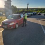 Police officers in high-visibility vests conduct a traffic stop at a roadside checkpoint; a red car is stopped in the foreground under a clear sky.