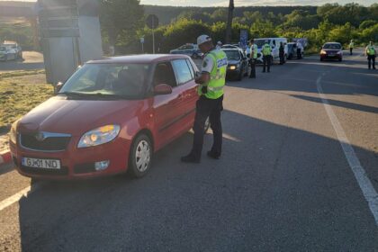 Police officers in high-visibility vests conduct a traffic stop at a roadside checkpoint; a red car is stopped in the foreground under a clear sky.