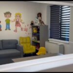 Playroom with a gray sofa, bright yellow chairs, and a cartoon wall mural of children and a dog, plus toy shelves in the corner.