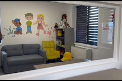Playroom with a gray sofa, bright yellow chairs, and a cartoon wall mural of children and a dog, plus toy shelves in the corner.