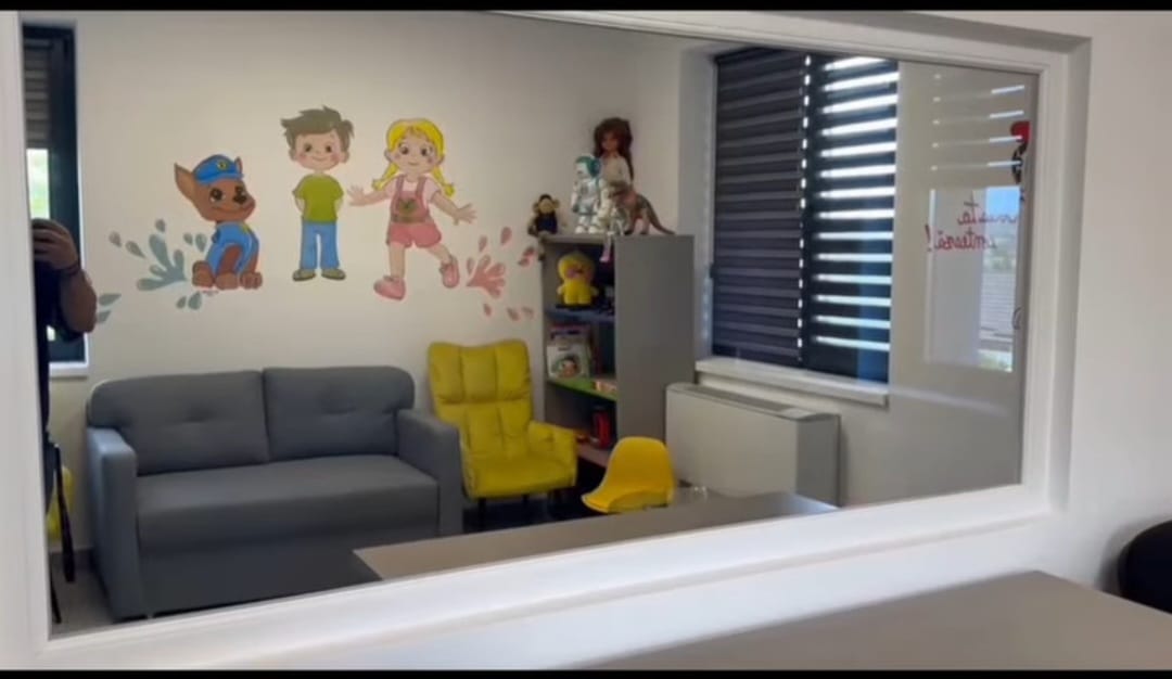 Playroom with a gray sofa, bright yellow chairs, and a cartoon wall mural of children and a dog, plus toy shelves in the corner.
