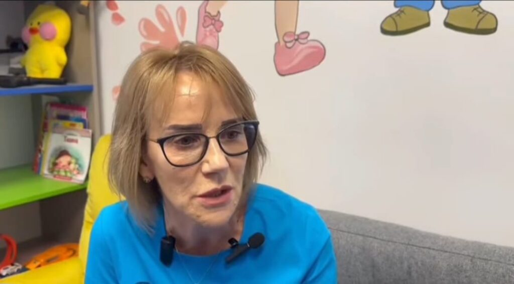 Mid-shot of a woman with short blond hair and glasses, wearing a blue top, speaking in a cozy room with colorful wall art and a yellow plush toy on a shelf in the background.