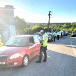 Police checkpoint on a sunlit road: an officer in a high‑visibility vest talks to the driver of a red car while other officers and police vehicles operate in the background on the roadside.