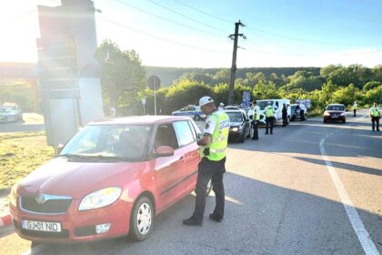 Police checkpoint on a sunlit road: an officer in a high‑visibility vest talks to the driver of a red car while other officers and police vehicles operate in the background on the roadside.