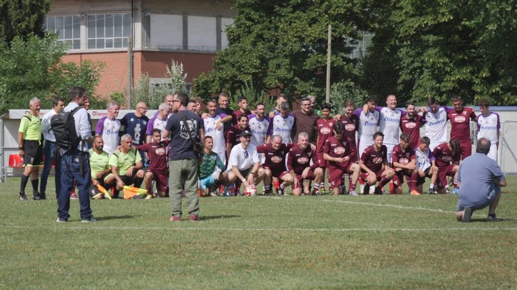 Group of soccer players and staff pose for a team photo on a grassy field; maroon and white uniforms, photographer kneeling to the right.