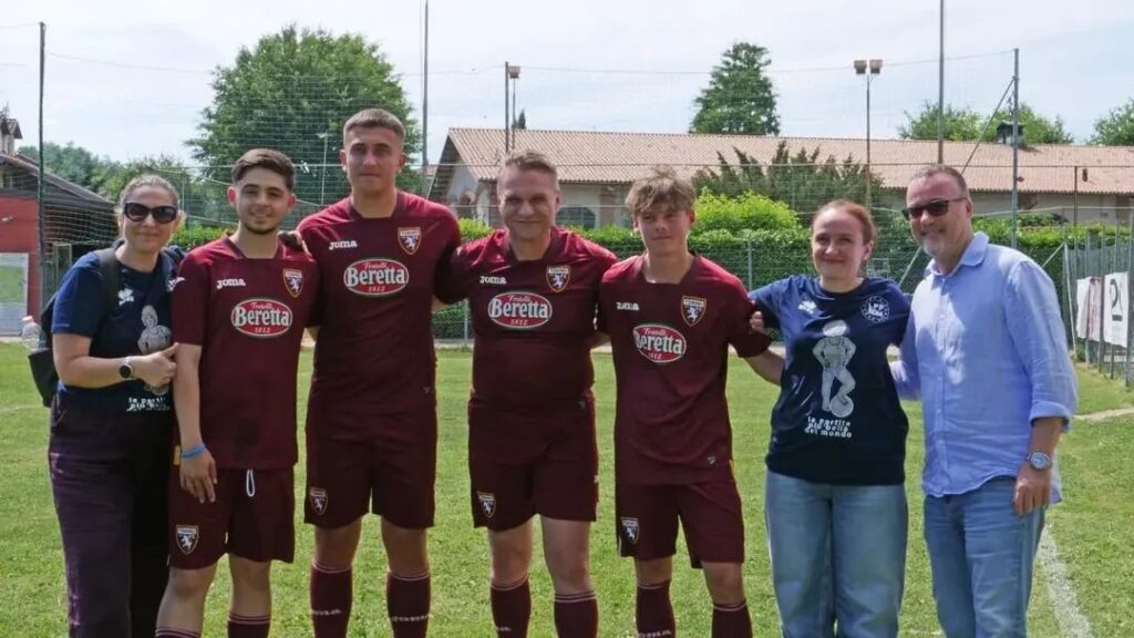 Group of seven people posing on a sunny soccer field; six players wear maroon kits with Beretta logo, flanked by two casually dressed adults on each end.
