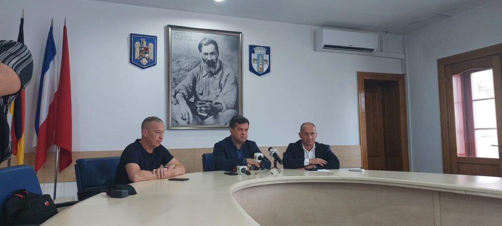Three men sit at a curved conference table with microphones in front of them during a press briefing, flags on the left and a large framed portrait on the wall behind.