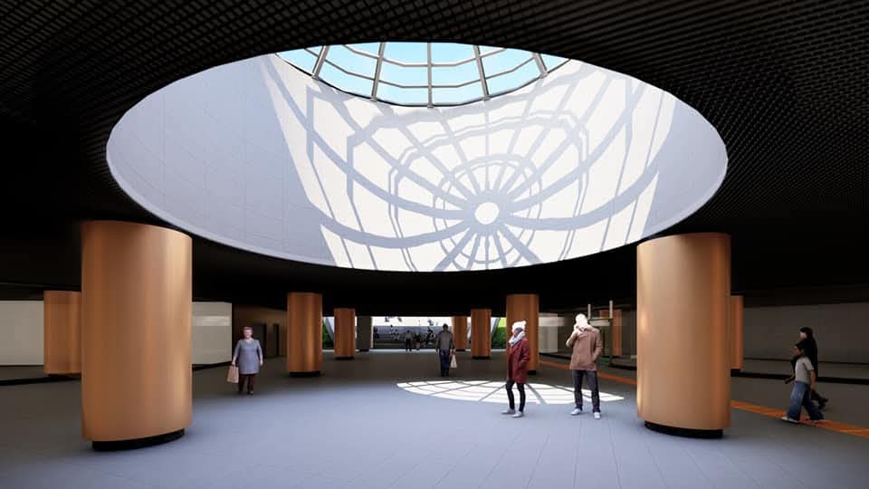 Underground transit hall with a large circular skylight casting geometric shadows on the floor; orange cylindrical columns support the space as people walk through.
