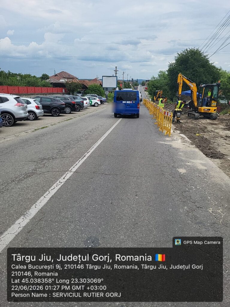 Road construction zone on a two-lane street with yellow barriers, an excavator, and workers beside the curb; blue van passing by.