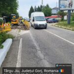 Road construction in progress: workers with yellow barriers and excavator along a cracked street as a white van passes by, Romania, Târgu Jiu.