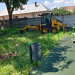 Yellow JCB compact excavator parked on overgrown grass beside a playground and fence behind a pink house.