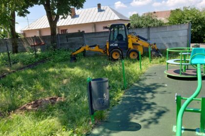 Yellow JCB compact excavator parked on overgrown grass beside a playground and fence behind a pink house.
