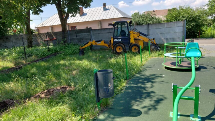 Yellow JCB compact excavator parked on overgrown grass beside a playground and fence behind a pink house.