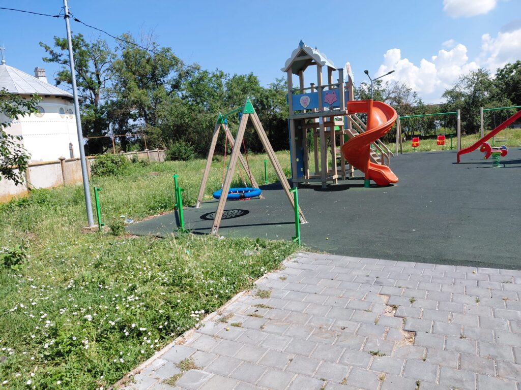 Playground with a wooden play structure, orange spiral slide and swings on a blacktop surface, surrounded by grass and trees under a clear blue sky. Nearby a white building is visible to the left.