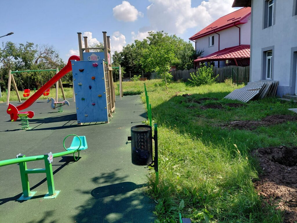 Playground with red slide, climbing wall, and swings on a sunny day, near a grassy yard and a white building with a red roof.