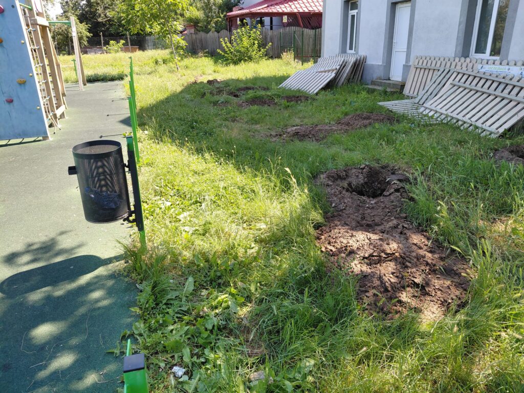 Playground area with a climbing wall on the left, green fencing, and a grassy, uneven yard near a gray building with stacked boards.