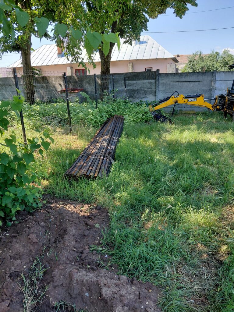 Construction site behind a fence with a yellow JCB excavator arm and a stack of metal beams on bare soil and grass.