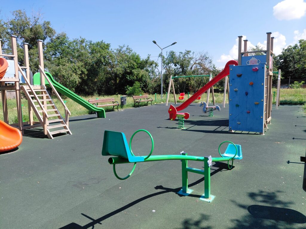 Children's playground with wooden play structures, green slide, red slide, and climbing wall on a sunny day.
