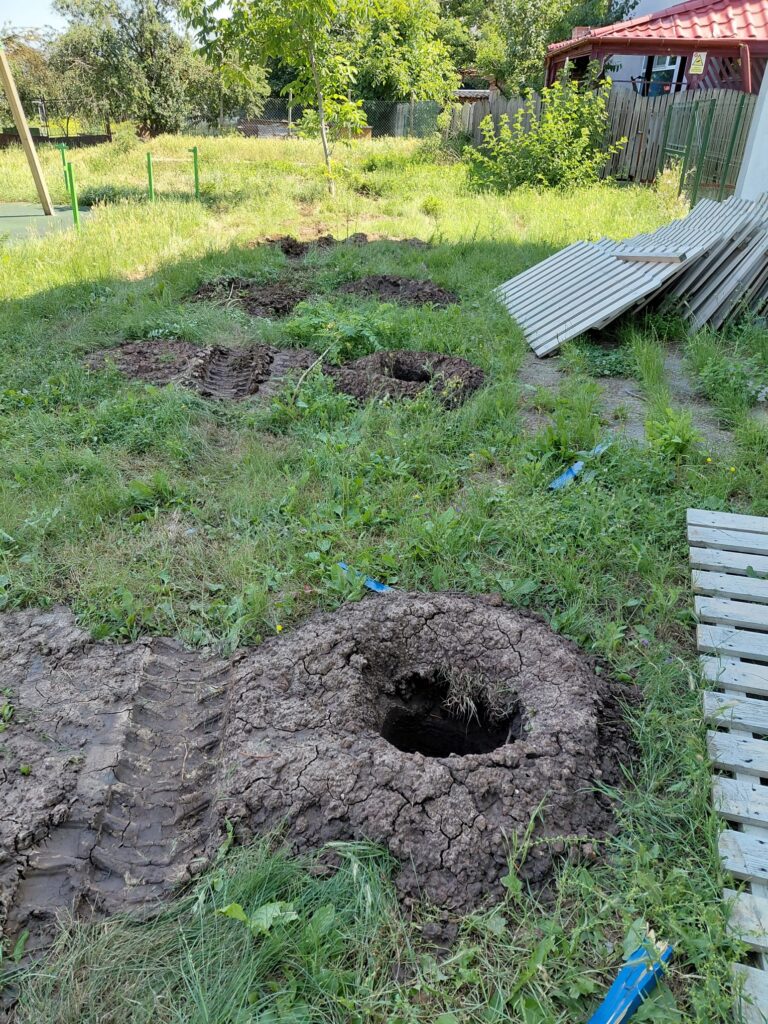Backyard with two large muddy holes in the ground, cracked earth around them, and corrugated metal sheets stacked on the right.
