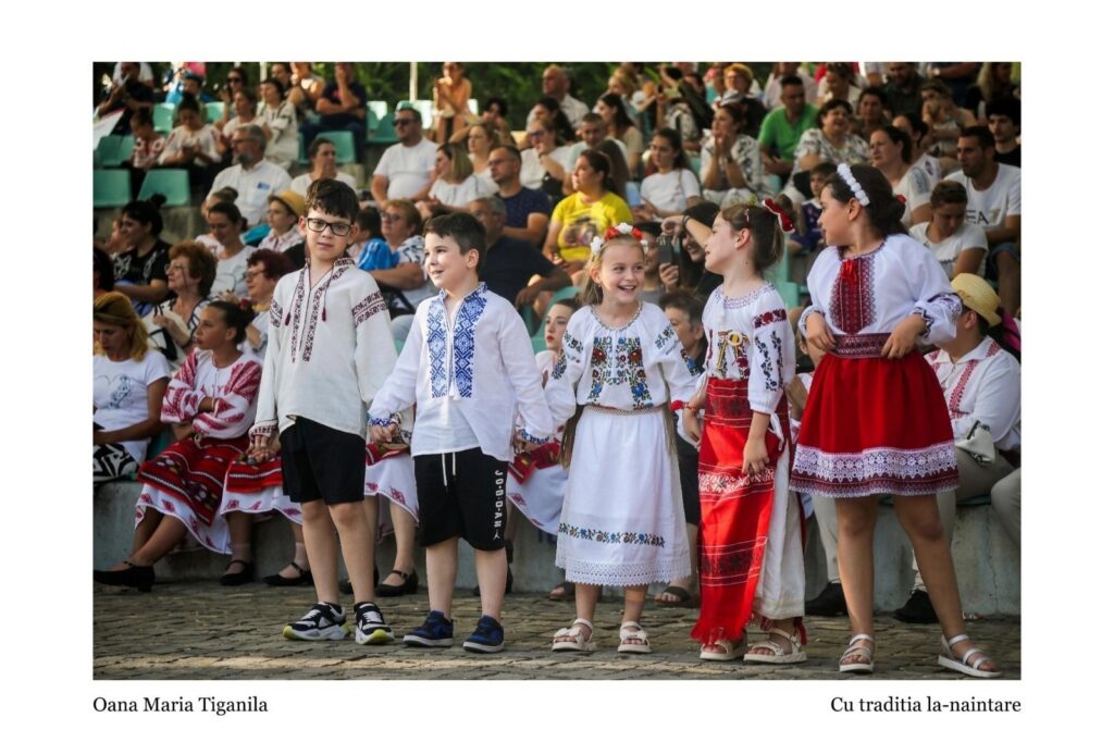 Children in embroidered folk costumes hold hands and walk in a line during a performance, with a seated audience in the background.