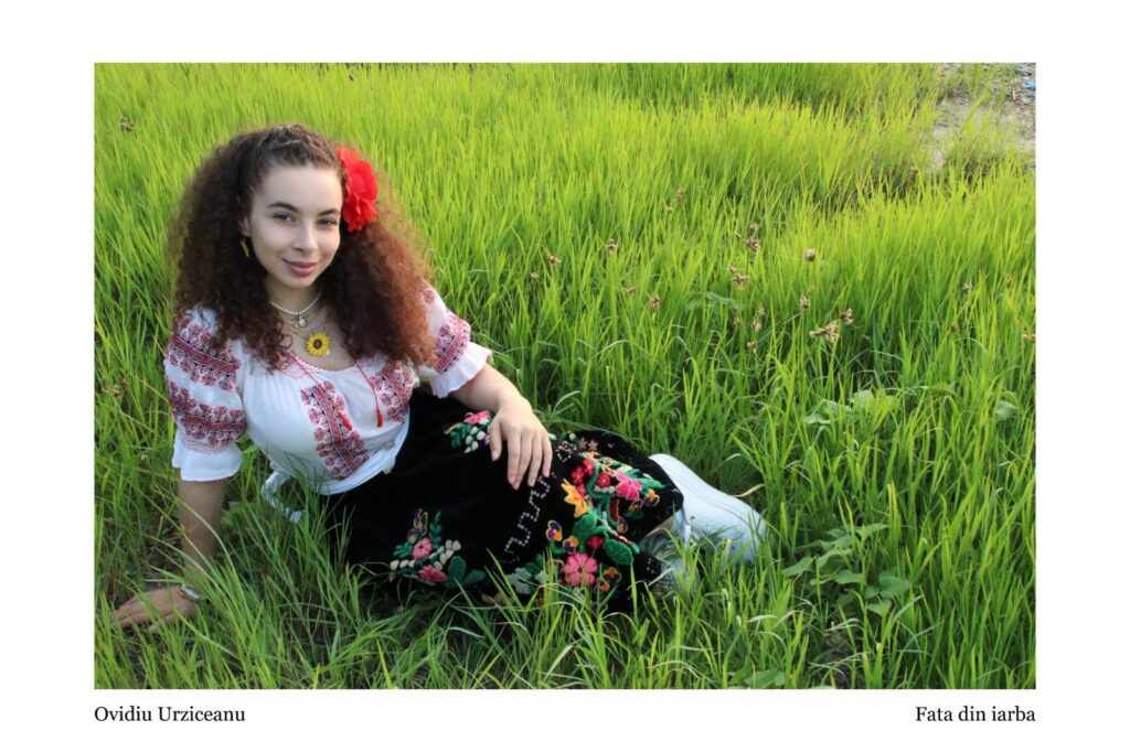 Portrait of a young woman with curly hair and a red flower in her hair, wearing a traditional embroidered blouse, sitting in a lush green field of tall grass.