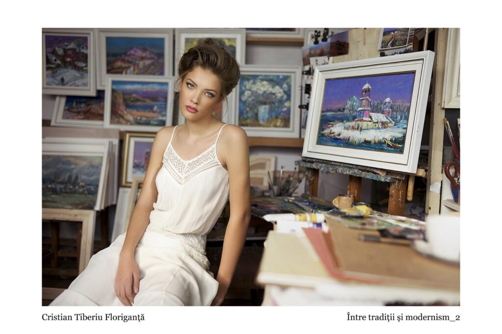 Portrait of a woman in a white lace-trim dress seated in an art studio, surrounded by framed paintings.
