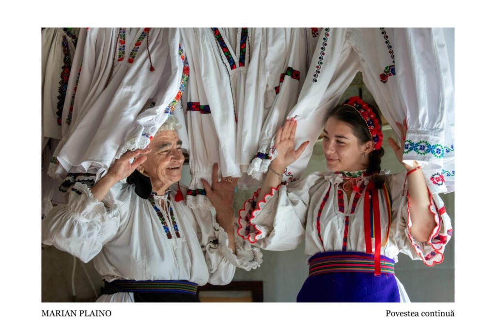 Two women in white folk costumes with colorful embroidery, smiling and raising hands under a canopy of embroidered shirts at a cultural event.