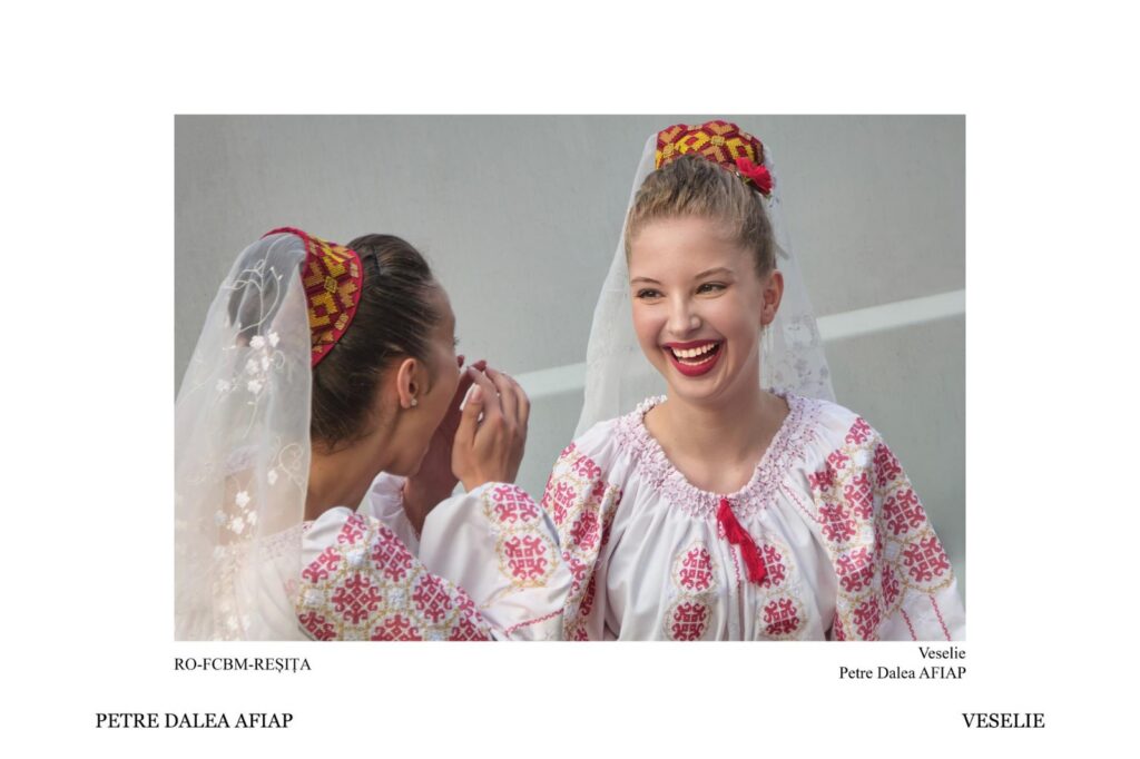 Two women in traditional white embroidered dresses with veils laugh together outdoors.