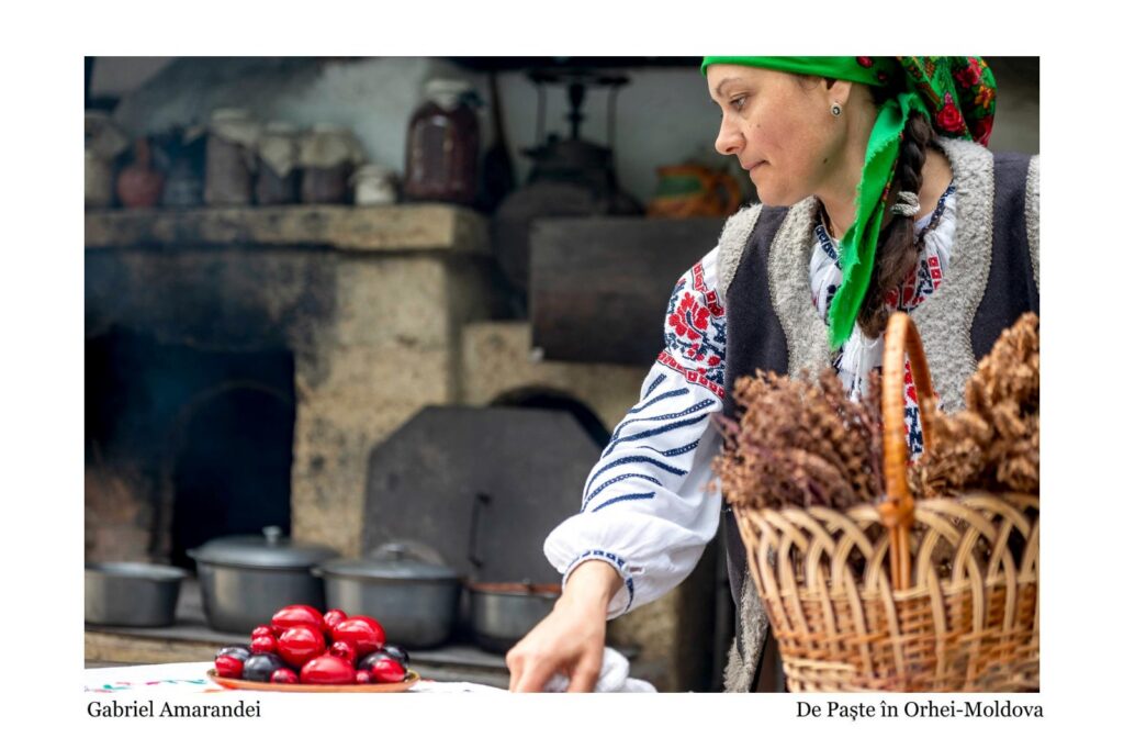 Traditional Romanian woman in folk clothing arranging red and black dyed eggs on a table, with a wicker basket nearby.