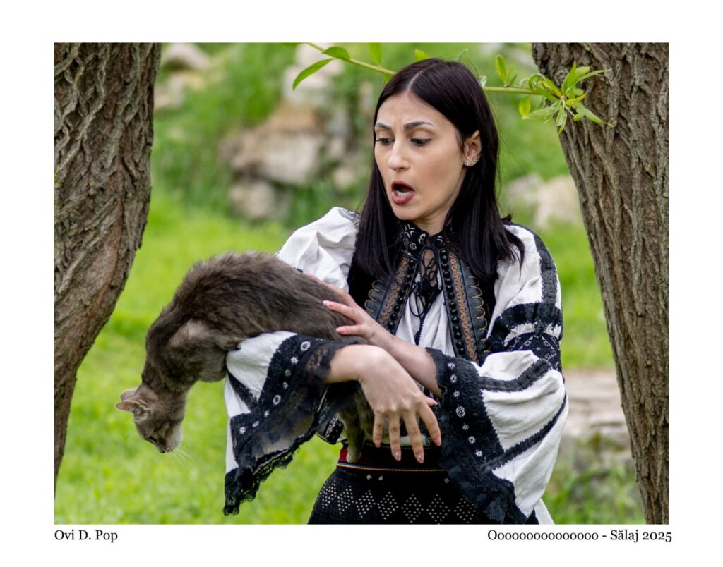 Woman in embroidered folk dress cradling a gray cat outdoors between two tree trunks.