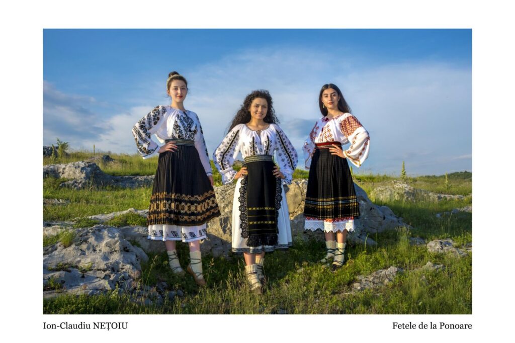 Three women in white embroidered blouses and black skirts pose on a rocky hill under a blue sky.