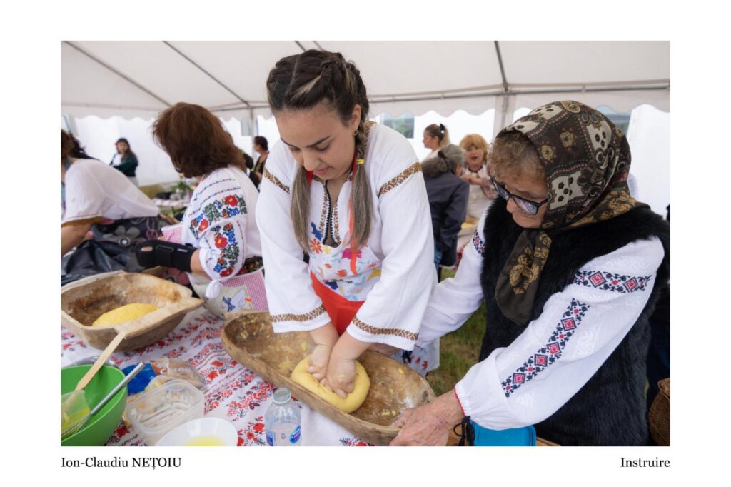 Women in traditional embroidered clothing kneading dough on a wooden trough at a festival under a white canopy