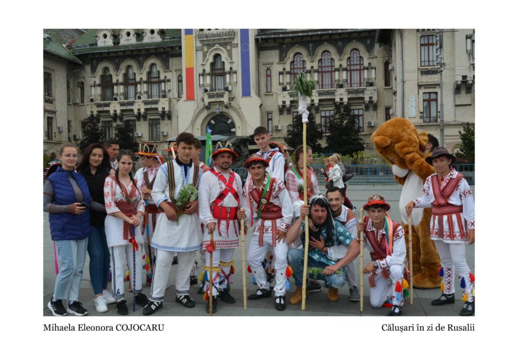 Group of people in colorful Romanian folk costumes posing for a festival photo in front of a historic building, with a bear costume in the background and onlookers nearby.