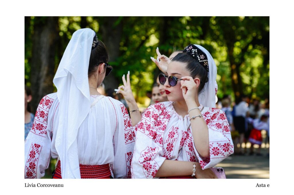 Two women in white dresses with red embroidery and headscarves talk to each other in a sunlit park, one wearing sunglasses and red lipstick.