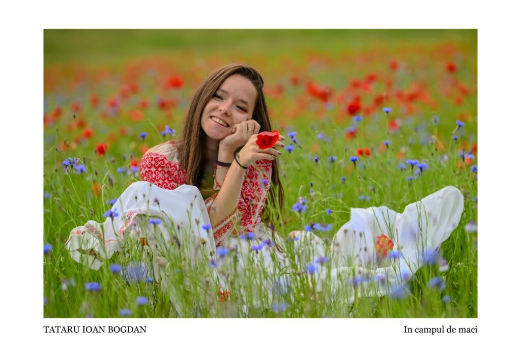Young woman in a red-embroidered dress sits in a field of red poppies and blue wildflowers, smiling at the camera.