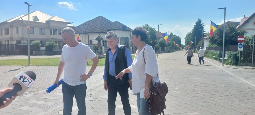 Three people stand on a sunny street during an interview, with a TV microphone visible in the foreground.