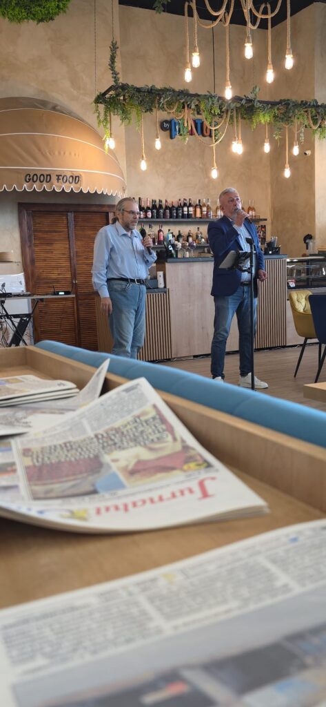Two men with microphones speak at a cafe bar, liquor bottles behind them and decorative hanging bulbs; newspapers in the foreground.