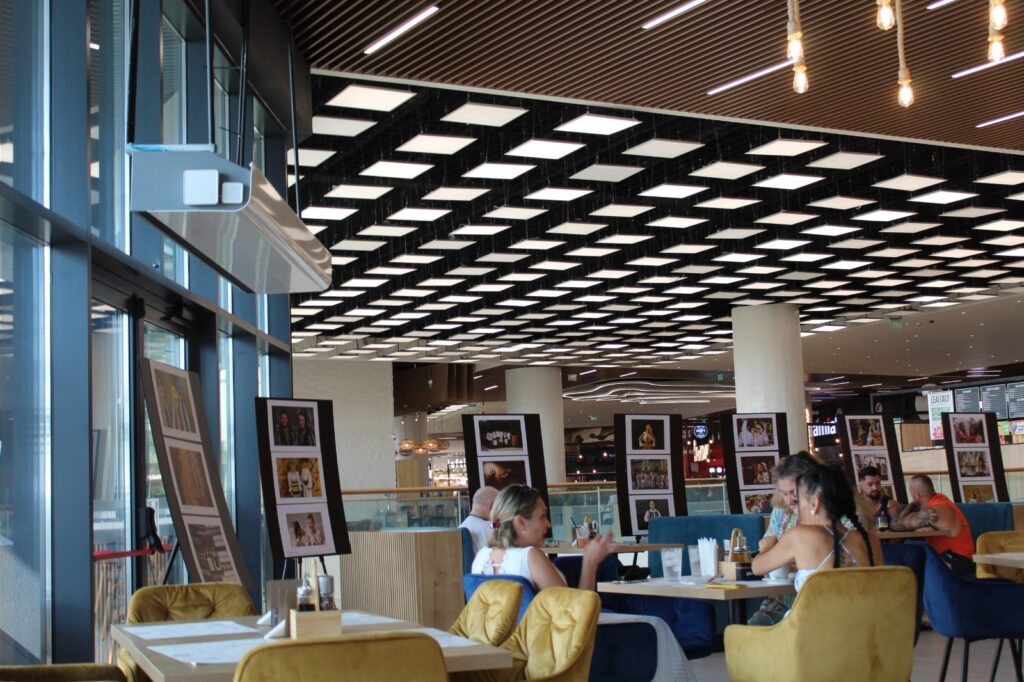 Modern cafe or food court with checkered white panel ceiling, diners at tables and framed photo displays nearby.