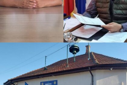 Man in a white T-shirt sits at a desk with hands folded, an office with flags behind him.”
