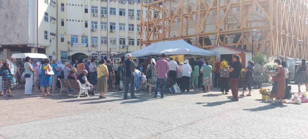 Crowd of people queuing at market stalls under a white canopy in a city square, with a wooden construction frame in the background.