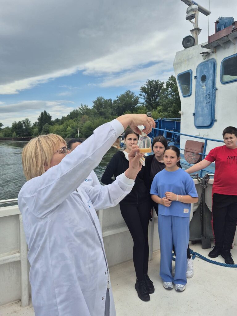 Scientist in a white lab coat showing a small bottle with yellow liquid to a group of kids on a boat deck with blue railings behind them.