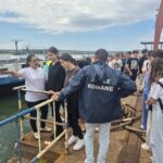 Group of students and a guide stand on a quay beside a moored boat at a port with a blue railing in the foreground.