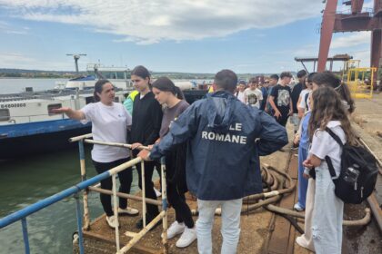 Group of students and a guide stand on a quay beside a moored boat at a port with a blue railing in the foreground.
