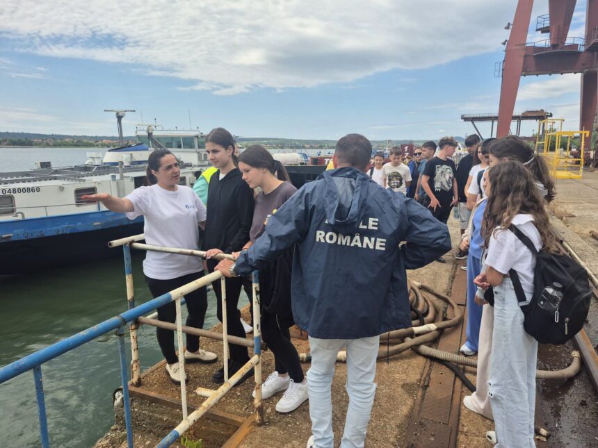 Group of students and a guide stand on a quay beside a moored boat at a port with a blue railing in the foreground.
