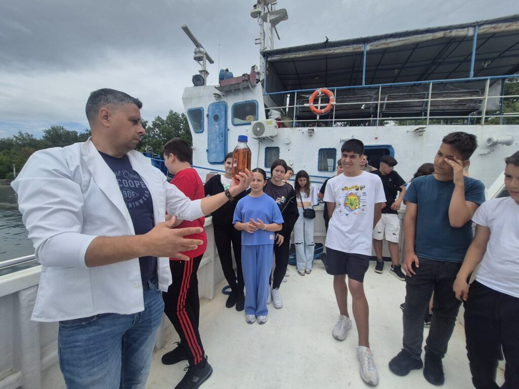 A man in a white lab coat gestures with a bottle while a group of teenagers stands on a boat deck and watches attentively, with a life ring and blue railings in the background.