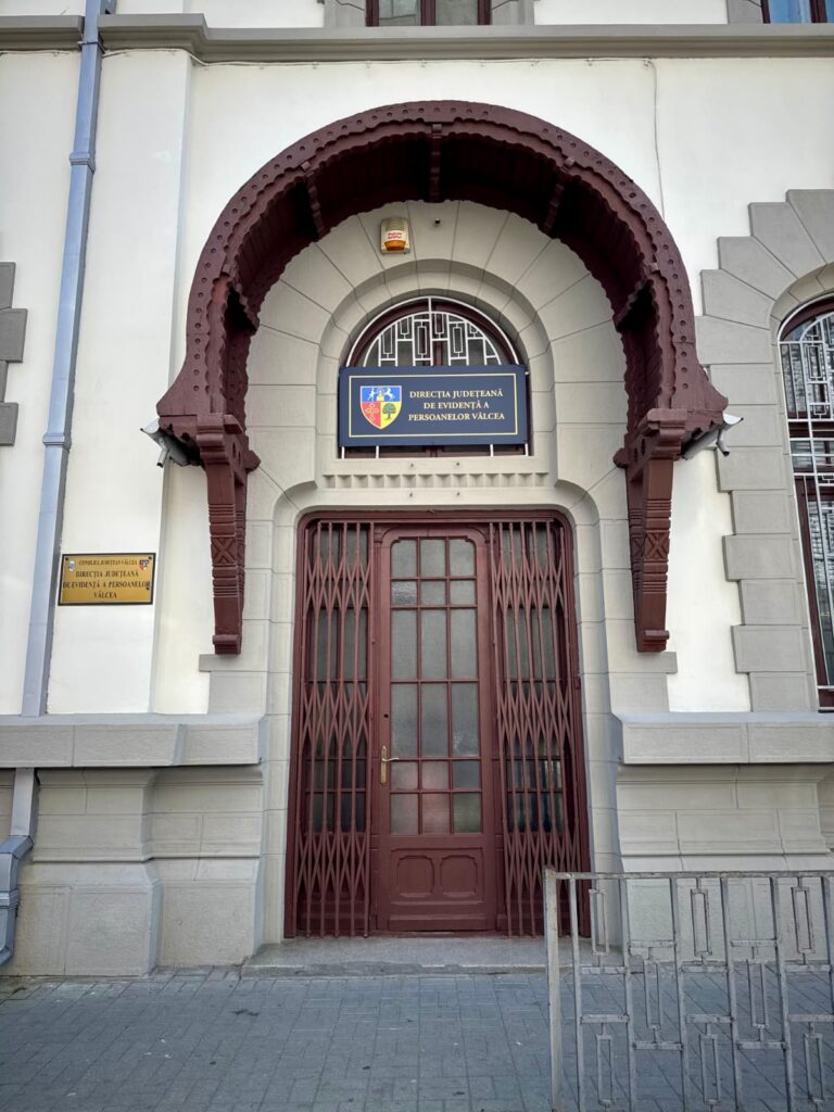 Entrance to a light-colored building with a large maroon rounded arch and iron gate; sign above reads Direcția Județeană de Evidență a Persoanelor Vâlcea.