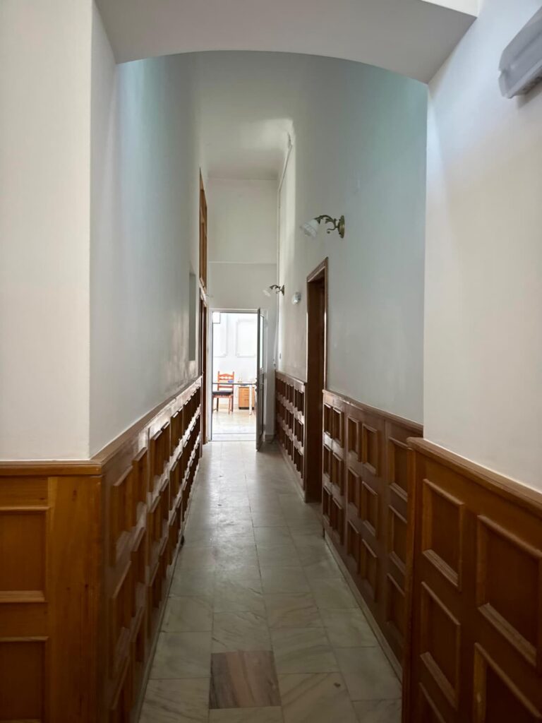 Narrow indoor corridor with light green walls, wooden wainscoting, and a marble floor leading to a sunlit room at the end with chairs visible through an open doorway.