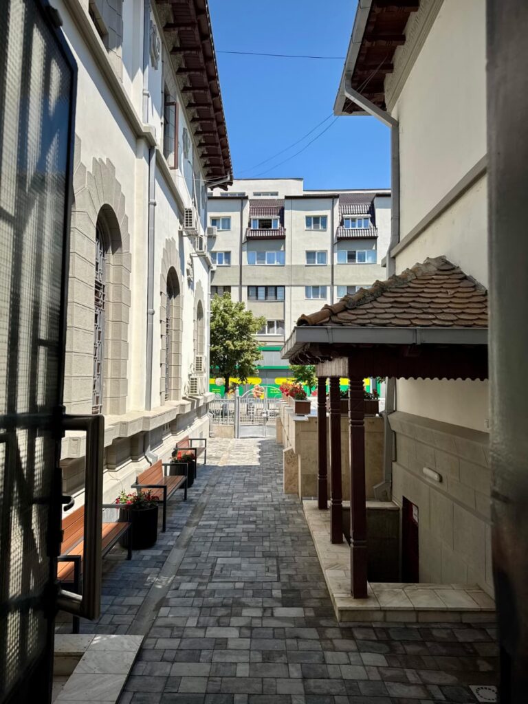 Narrow walkway between light historic buildings with arched windows and benches on the left, leading to a modern apartment building under a bright blue sky.